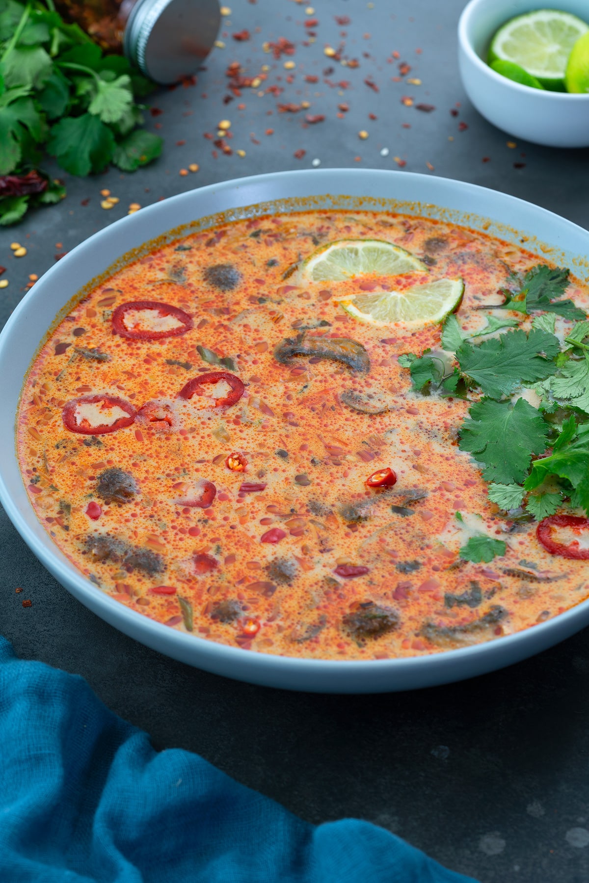 Thai Tom Kha Gai Soup in a white bowl with few ingredients scattered around. 