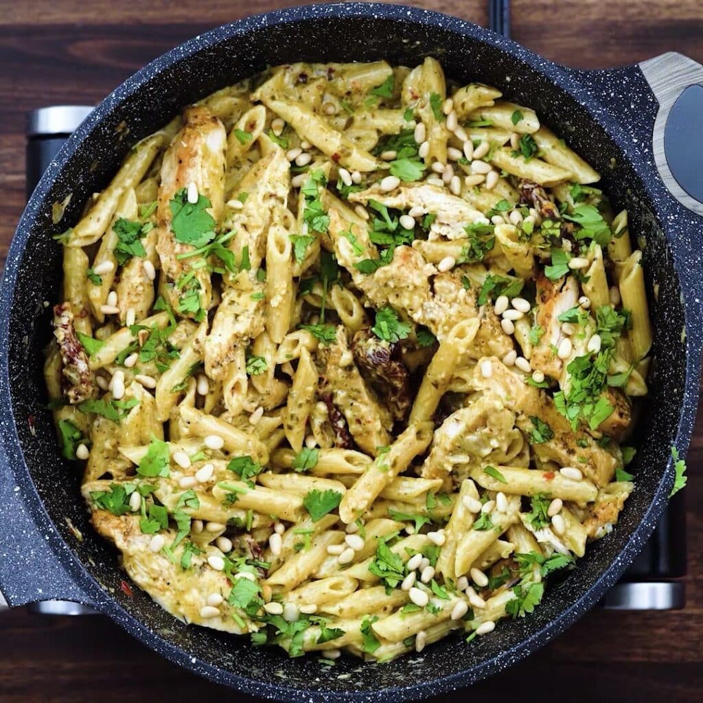 A pan with pesto chicken pasta garnished with herbs and nuts.