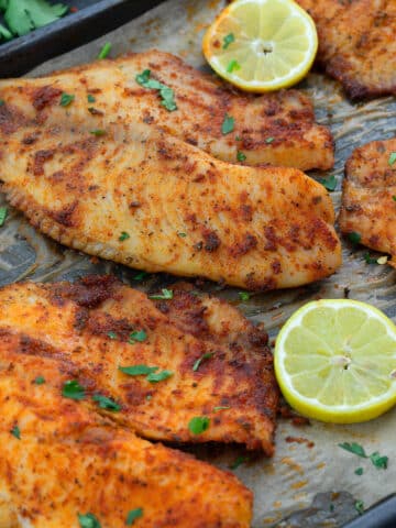 Oven Baked Tilapia in a baking tray with lemon slices, and few ingredients scattered around.