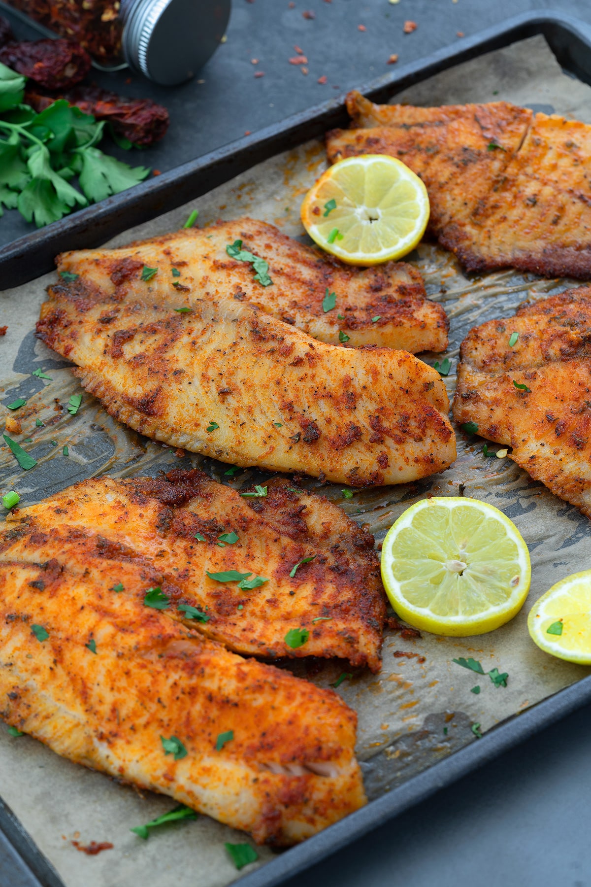Oven Baked Tilapia in a baking tray with lemon slices, and few ingredients scattered around.