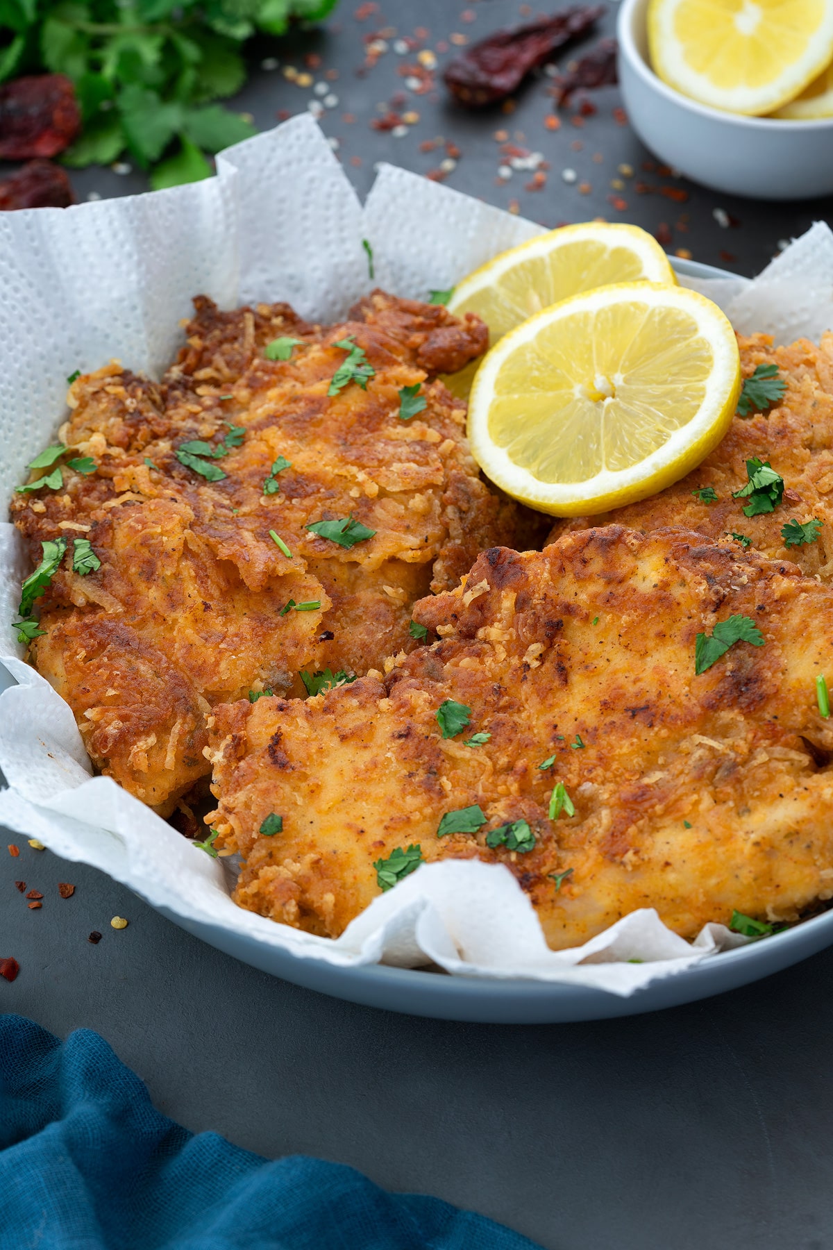 Parmesan Crusted Chicken in a bowl with lemon slides with few ingredients scattered around.