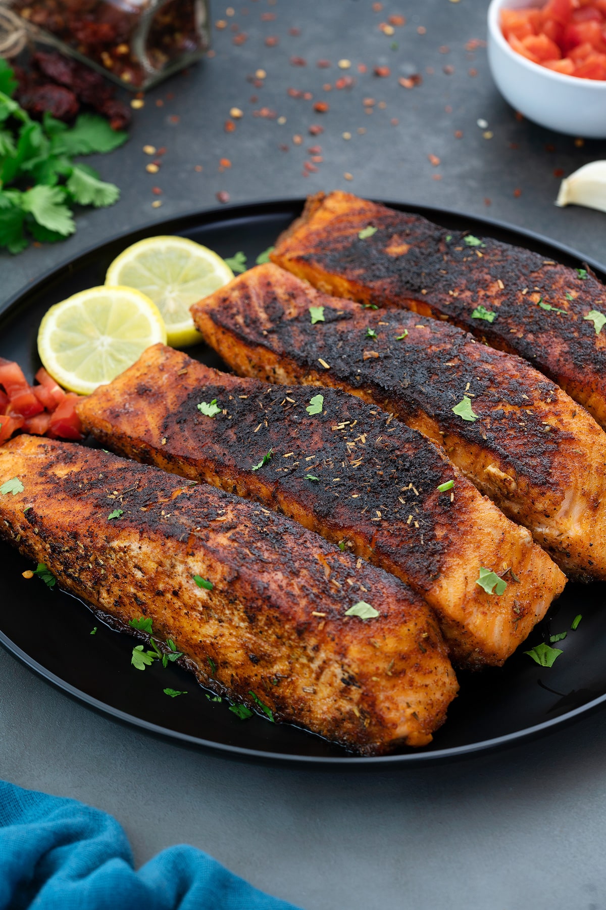 Homemade Blackened Salmon in a black plate with lemon slices and tomato pieces with few ingredients scattered around.