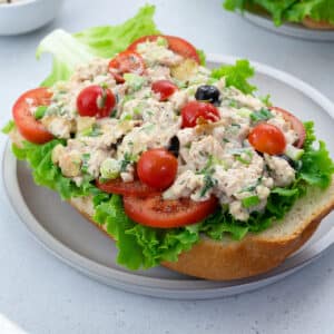 Chicken Salad with cherry tomatoes, lettuce, and tomato slices on bread. Served on a white plate on a white table.