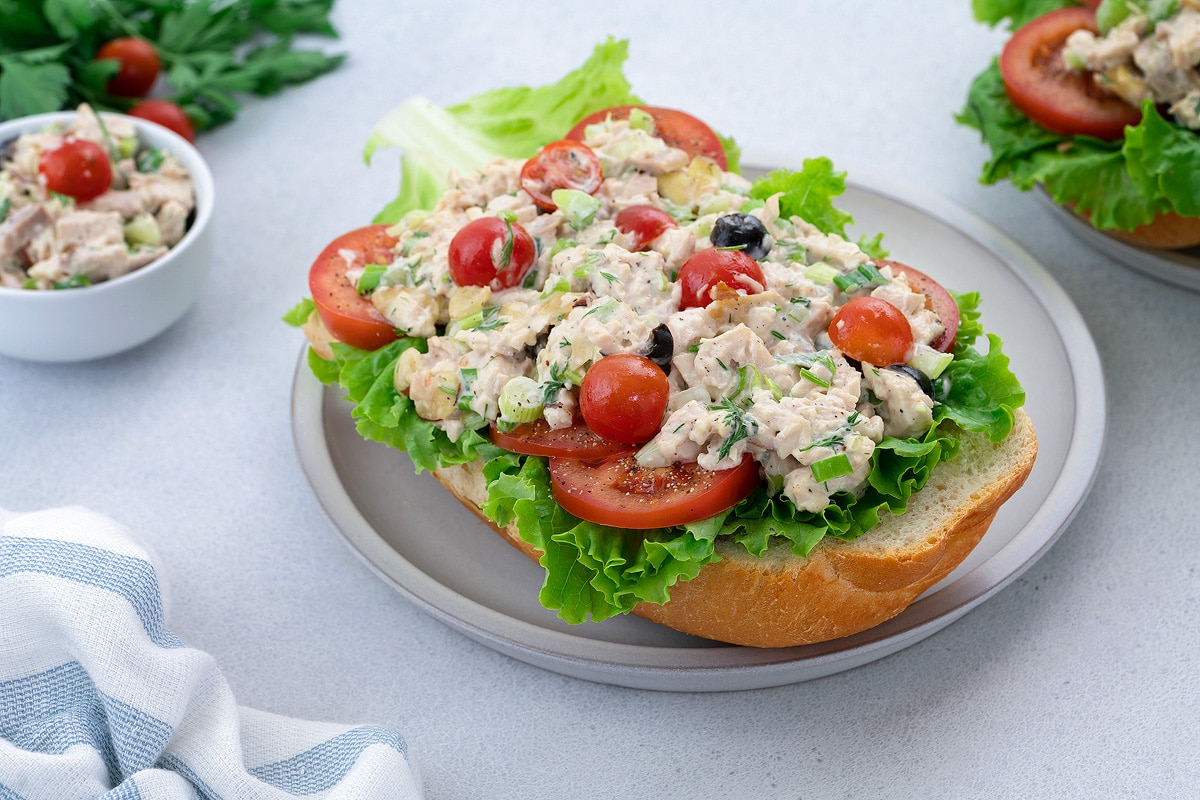 Chicken Salad with cherry tomatoes, lettuce, and tomato slices on bread. Served on a white plate on a white table.