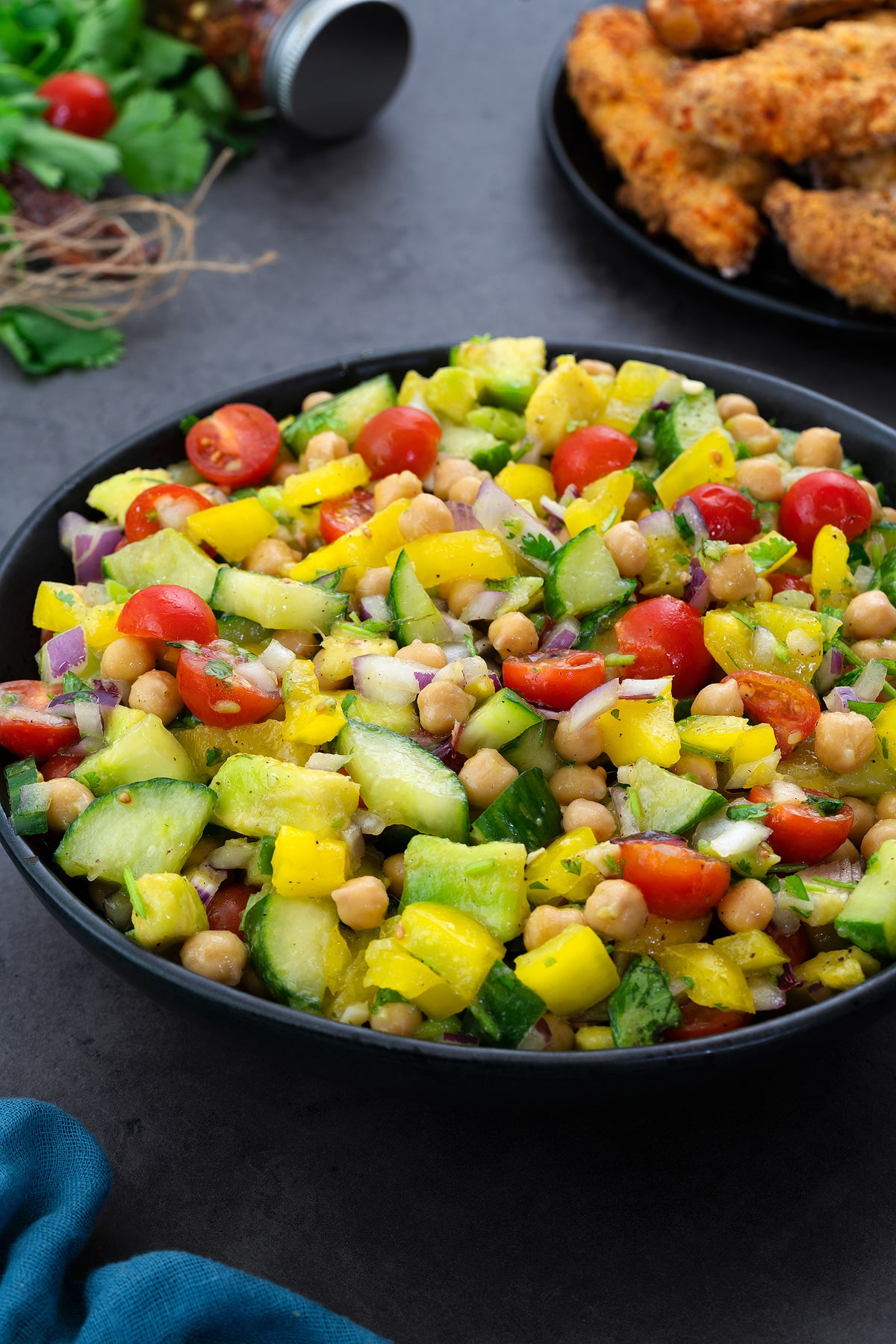 Chickpea salad in a black bowl on a grey table.  The salad is green, red and yellow, and it is surrounded by pieces of chicken and greens.