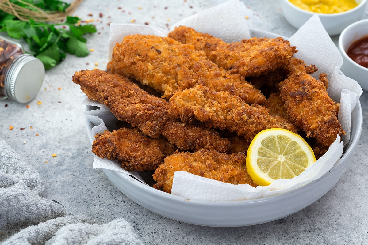 Fried Chicken Tenders in a white bowl on a white table with few other ingredients arranged around.