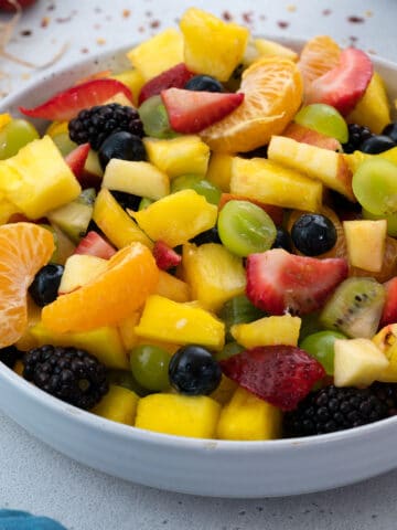 Homemade fruit salad with a variety of fruits in a white bowl placed on a clean white table.