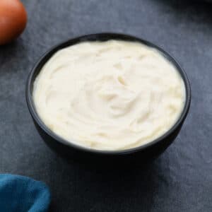 Homemade Mayonnaise in a black bowl placed on a grey table with an egg nearby.
