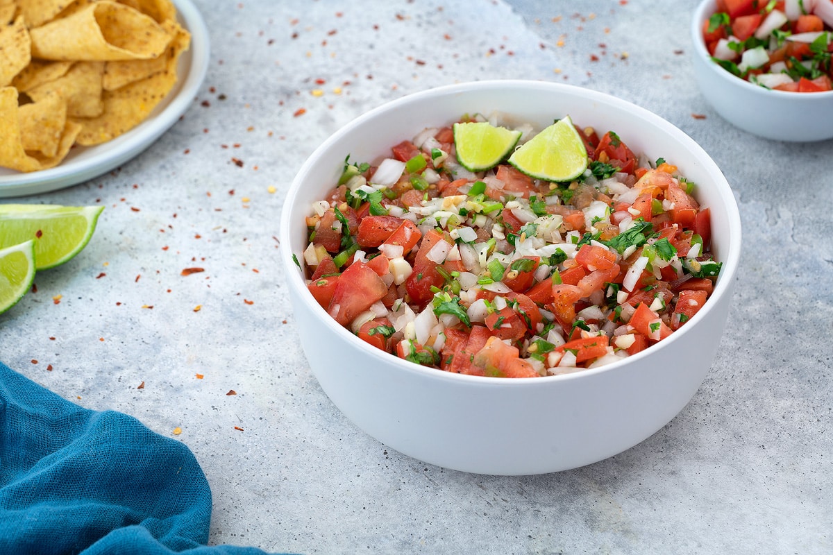 A bowl of pico de gallo with fresh tomatoes, onion, jalapeno, lime, and cilantro on a white table.