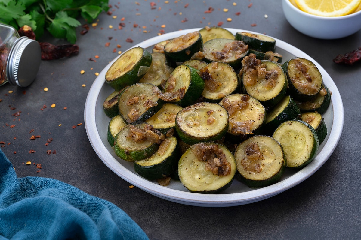 Sauteed zucchini in a white bowl on a gray table. The dish includes zucchini, onions, and garlic.