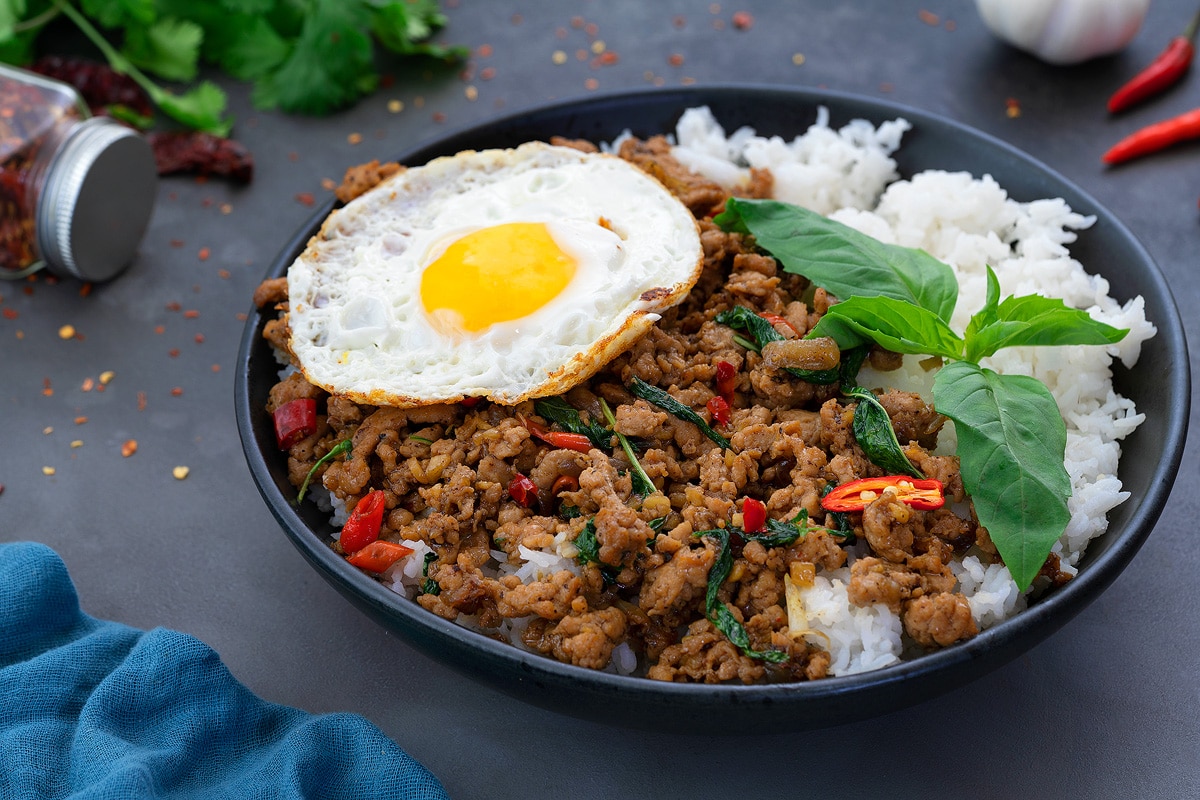 Thai Basil Chicken served in a black bowl, accompanied by fried eggs and rice. Various ingredients are scattered around the dish.