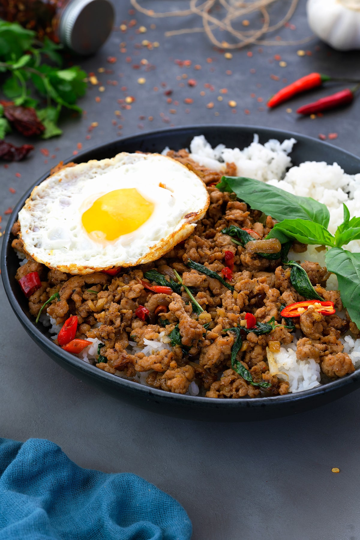 Thai Basil Chicken served in a black bowl, accompanied by fried eggs and rice. Various ingredients are scattered around the dish.