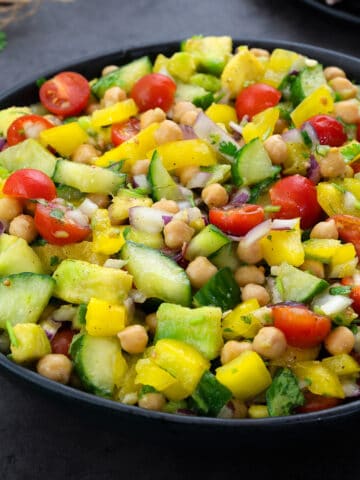 Chickpea salad in a black bowl on a grey table. The salad is green, red and yellow, and it is surrounded by pieces of chicken and greens.