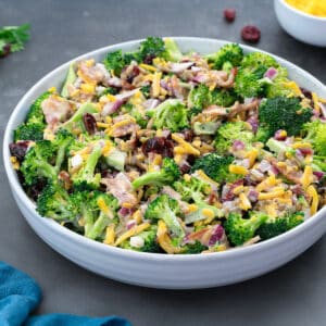 Close up of Broccoli salad in a white bowl on a grey table, made with broccoli, cheddar cheese, sunflower seeds, bacon, cranberries, and mayo. Additional cheddar cheese and greens on the table.