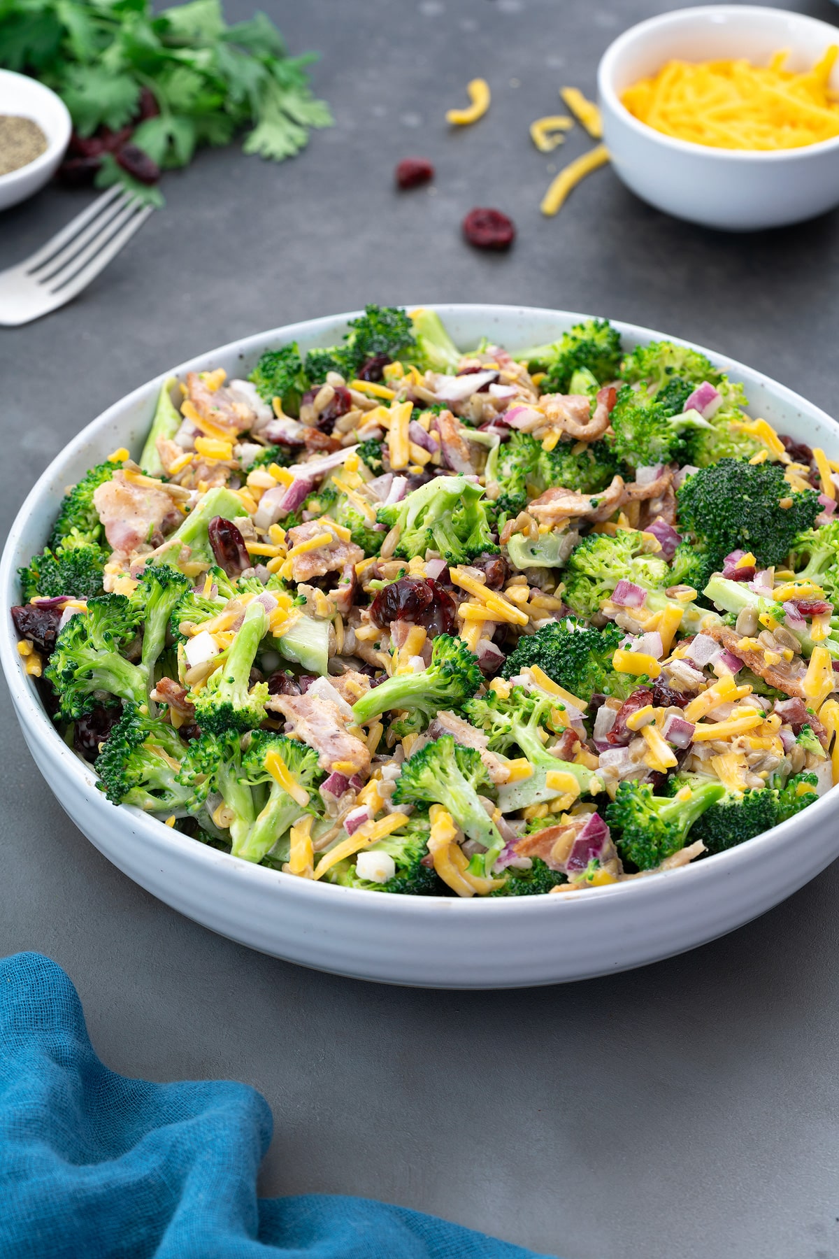 Broccoli salad in a white bowl on a grey table, made with broccoli, cheddar cheese, sunflower seeds, bacon, cranberries, and mayo. Additional cheddar cheese and greens on the table.