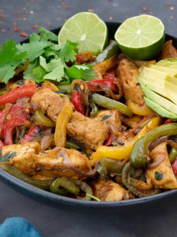 Vibrant Homemade Chicken Fajitas showcased in a black bowl on a grey table. The dish bursts with the rich hues of green, yellow, and red bell peppers, complemented by fresh greens and a zesty lime slices.