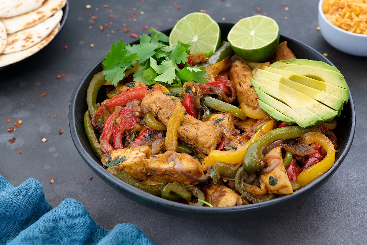 Vibrant Homemade Chicken Fajitas showcased in a black bowl on a grey table. The dish bursts with the rich hues of green, yellow, and red bell peppers, complemented by fresh greens and a zesty lime slices.