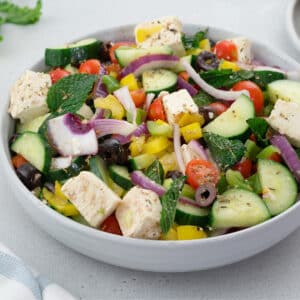 A colorful Greek Salad made of cucumber, green and red bell pepper, feta cheese, kalamata olives, and mint leaves. The salad is placed in a white bowl on a white table.