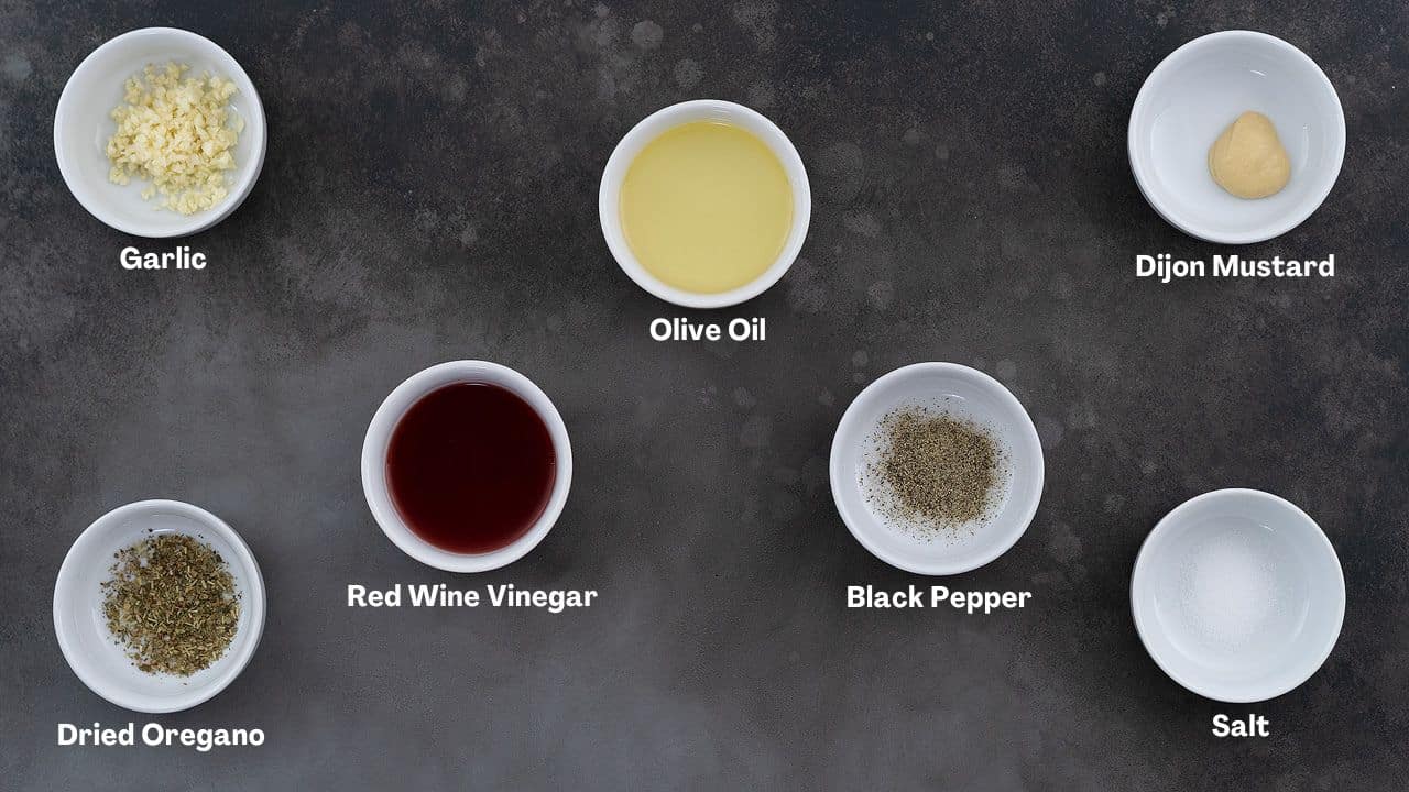 Ingredients for Greek Salad dressing in cups, including red wine vinegar, oregano, Dijon mustard, garlic, and pepper powder, arranged on a gray table.