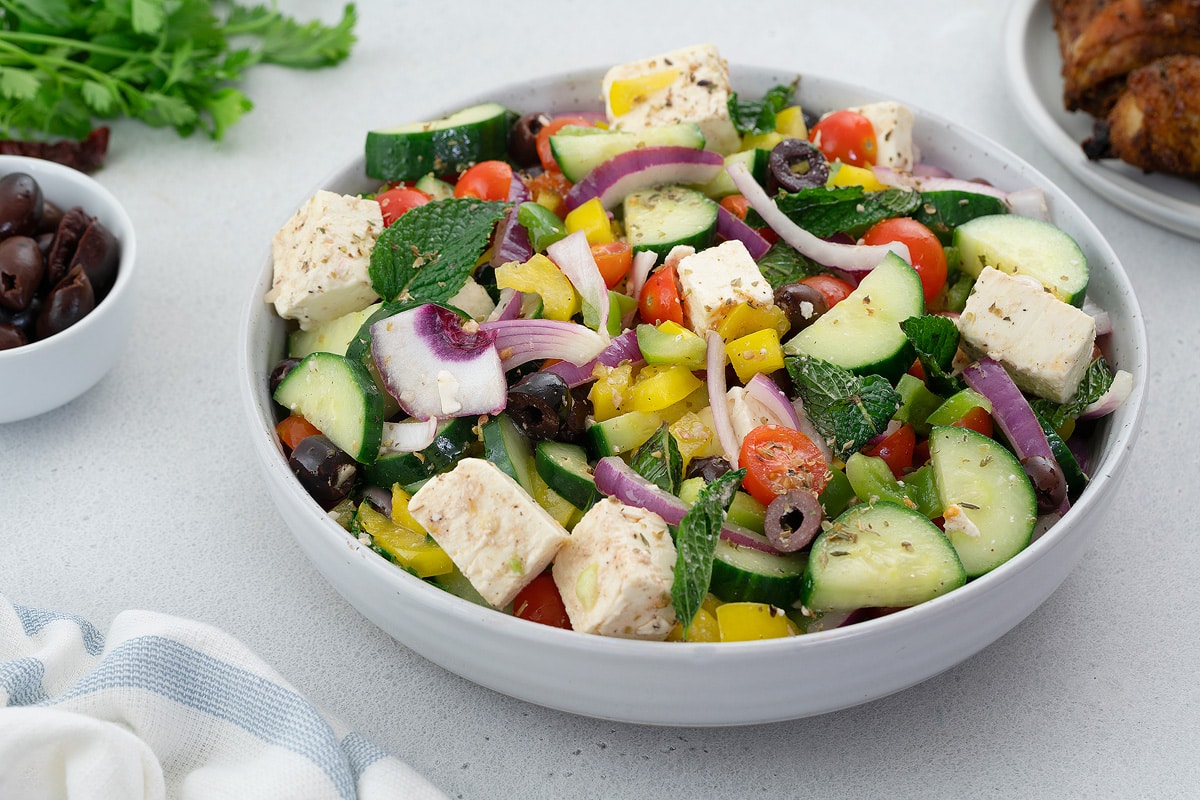 A colorful Greek Salad made of cucumber, green and red bell pepper, feta cheese, kalamata olives, and mint leaves. The salad is placed in a white bowl on a white table. There are also chicken and olives in a plate and a cup.