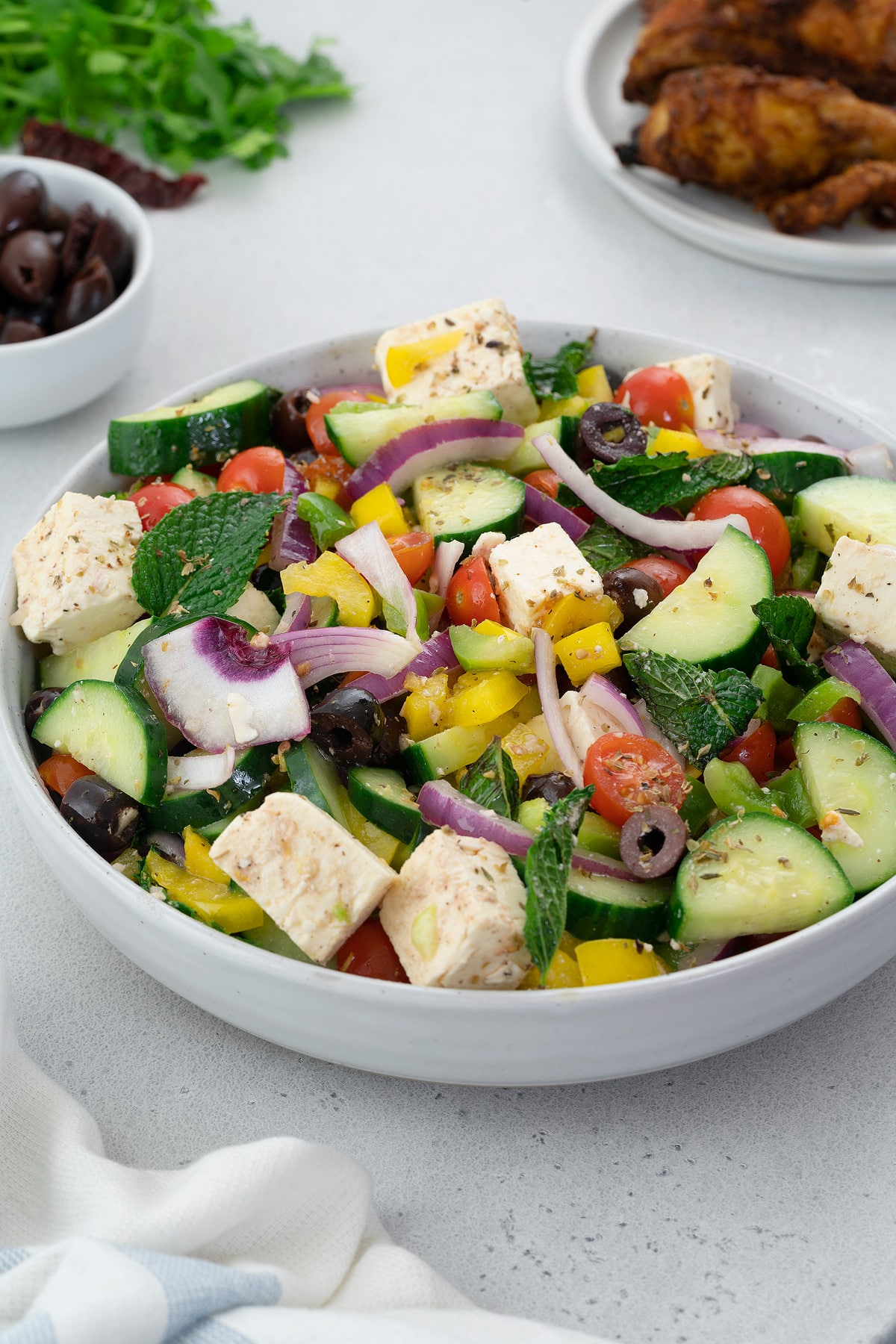 A colorful Greek Salad made of cucumber, green and red bell pepper, feta cheese, kalamata olives, and mint leaves. The salad is placed in a white bowl on a white table. There are also chicken and olives in a plate and a cup.