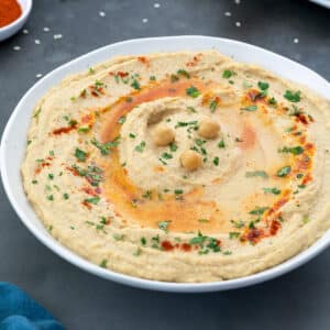 Close-up of homemade hummus in a white bowl placed on a grey table, garnished with olive oil, paprika, and coriander leaves.