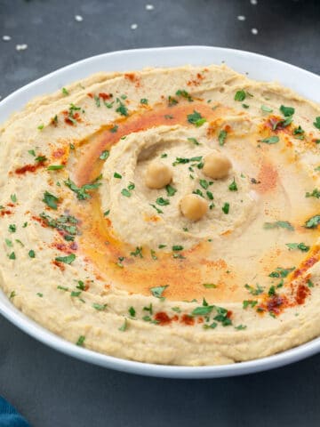 Close-up of homemade hummus in a white bowl placed on a grey table, garnished with olive oil, paprika, and coriander leaves.
