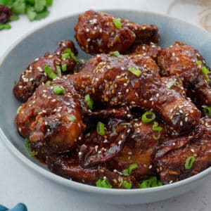 Close up of Homemade Korean fried chicken (drumsticks, wings) in a light blue bowl on a white table. The chicken is golden brown and coated in a sweet and spicy sauce.