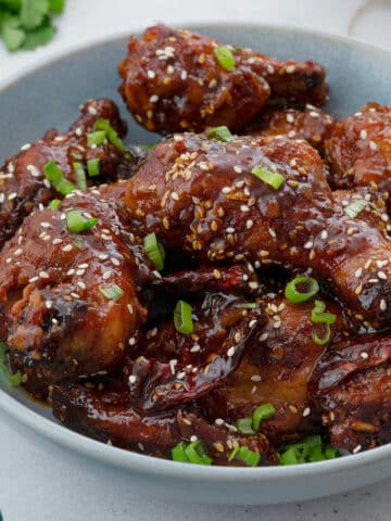 Close up of Homemade Korean fried chicken (drumsticks, wings) in a light blue bowl on a white table. The chicken is golden brown and coated in a sweet and spicy sauce.