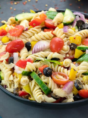 Homemade Pasta Salad in a black bowl on a grey table, featuring Mozzarella Cheese, Pasta, Grape Tomatoes, Bell Pepper, Cucumber, Basil Leaves, and other ingredients.