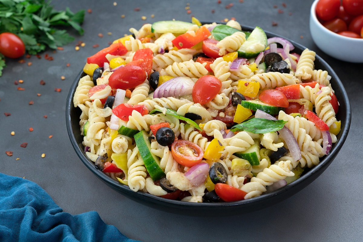 Homemade Pasta Salad in a black bowl on a grey table, featuring Mozzarella Cheese, Pasta, Grape Tomatoes, Bell Pepper, Cucumber, Basil Leaves, and other ingredients. The dish is accompanied by a cup of grape tomatoes and garnished with greens.