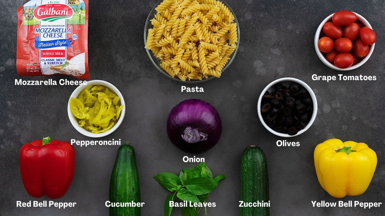 Pasta Salad recipe Ingredients on a grey table featuring Mozzarella Cheese, Pasta, Grape Tomatoes, Bell Pepper, Cucumber, Basil Leaves, and other ingredients.