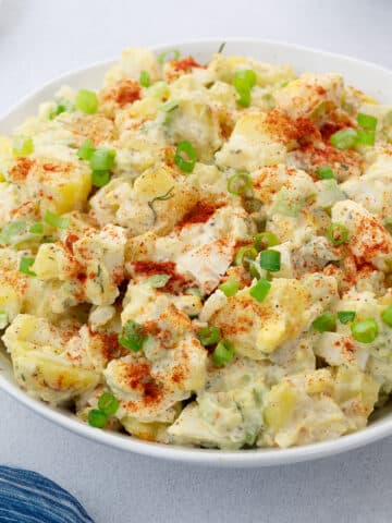 Close up of Homemade Potato Salad in a white bowl, garnished with a sprinkle of paprika and fresh spring onions, served on a white table.