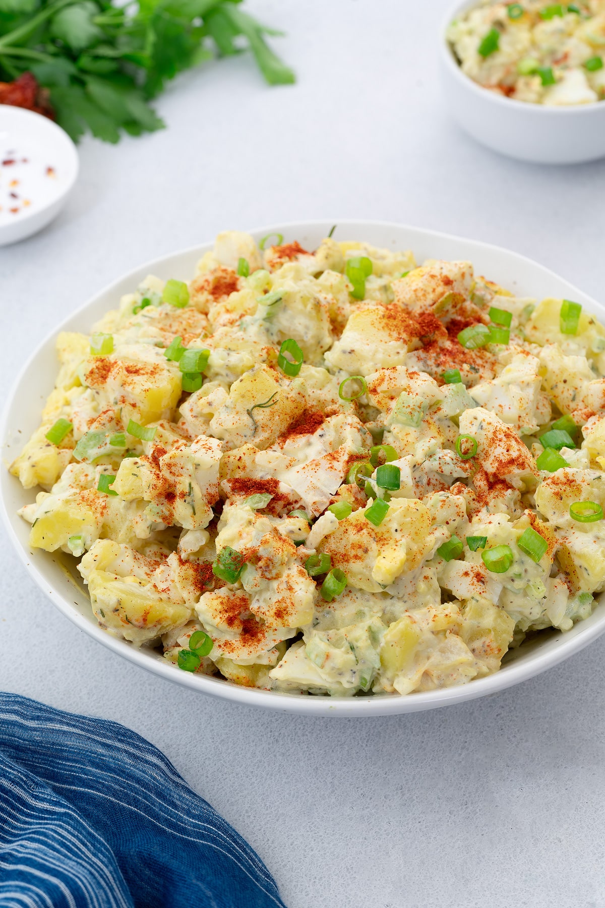 Delicious Homemade Potato Salad in a white bowl, garnished with a sprinkle of paprika and fresh spring onions, served on a white table.