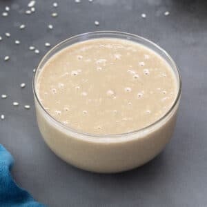Close up of homemade tahini in a clear glass bowl on a grey table with a blue color towel in the buttom left corner. And sesame seeds are scattered around the bowl.