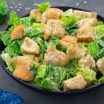 Homemade Caesar salad in a black bowl on a grey table. A cup of Caesar salad dressing and a Romaine lettuce leaf placed nearby.