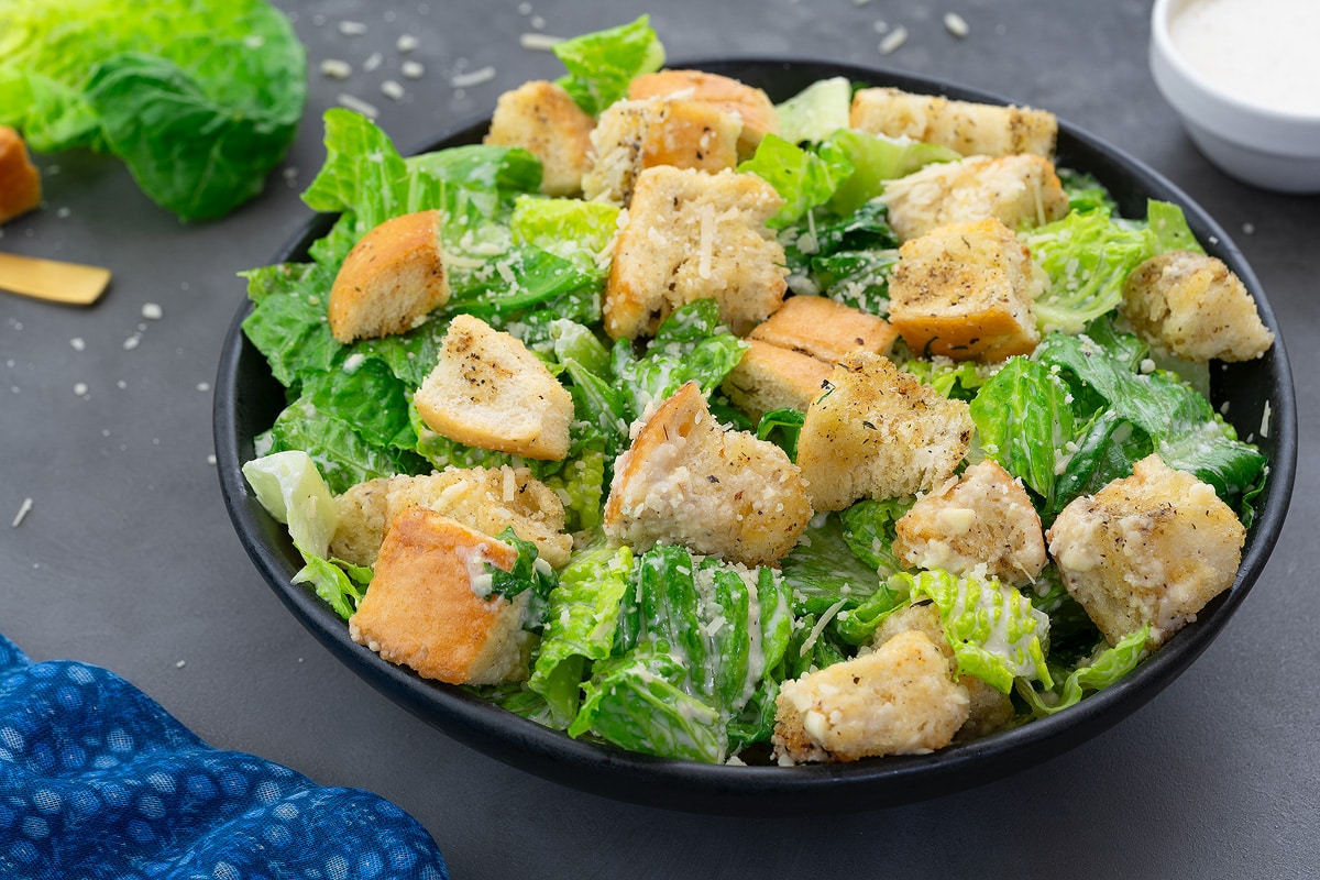 Homemade Caesar salad in a black bowl on a grey table. A cup of Caesar salad dressing and a Romaine lettuce leaf placed nearby.