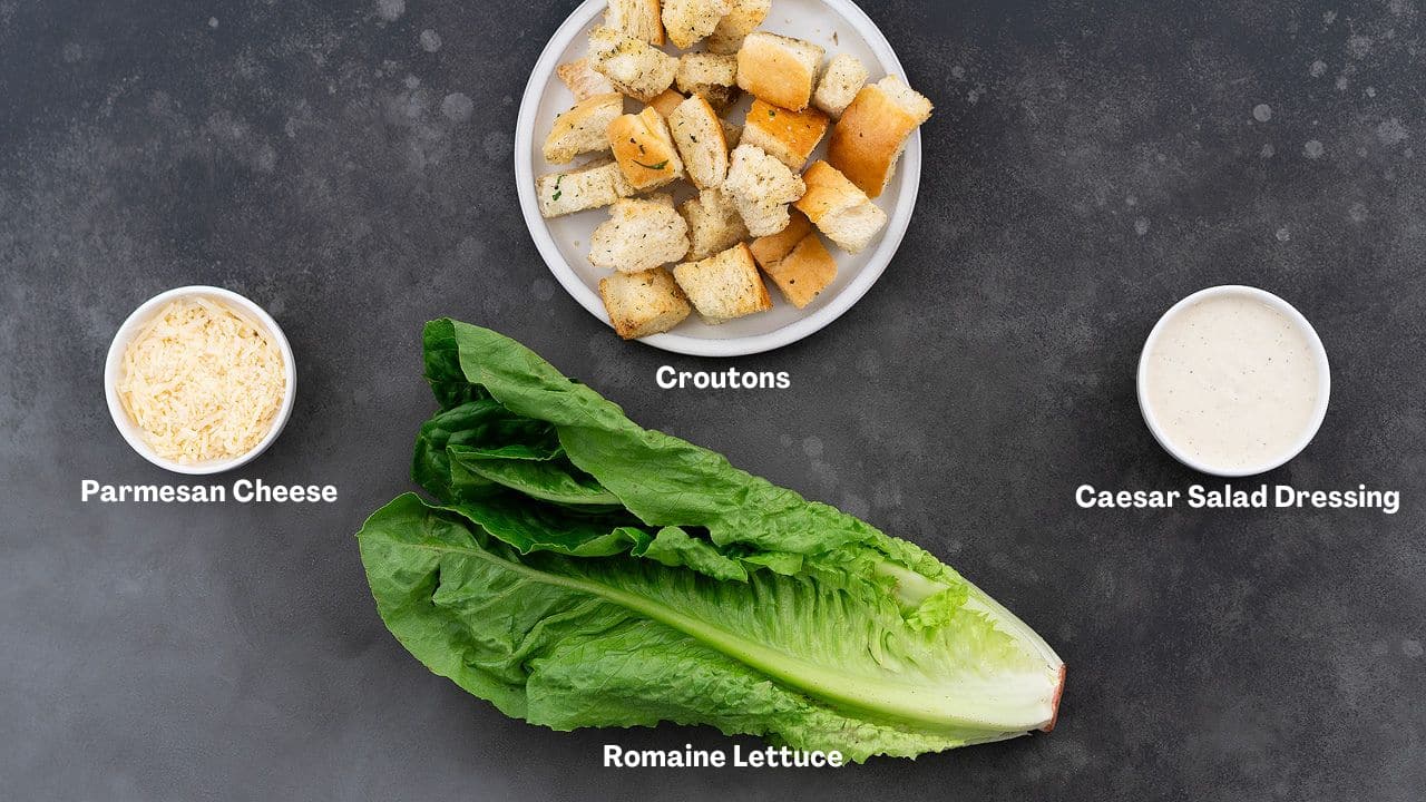 Caesar salad ingredients arranged on a grey table: croutons, Parmesan cheese, Romaine lettuce, and Caesar salad dressing.