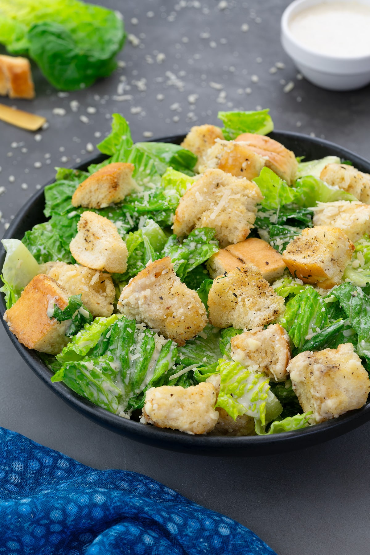 Homemade Caesar salad in a black bowl on a grey table. A cup of Caesar salad dressing and a Romaine lettuce leaf placed nearby.