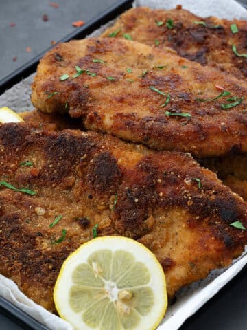 Close up of crispy and tender Chicken Cutlets on a black tray, presented on a grey table with fresh coriander leaves and zesty lemon slices.