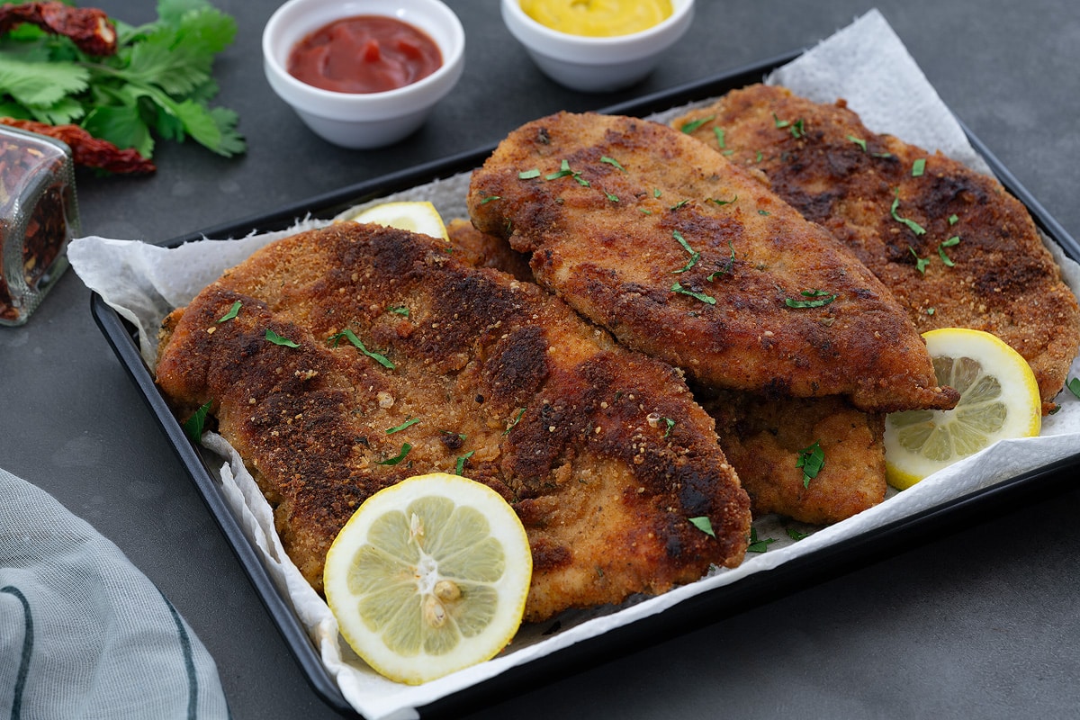 Crispy and Tender Chicken Cutlets on a black tray, presented on a grey table with fresh coriander leaves and zesty lemon slices. Accompanied by white cups of flavorful Ketchup and yellow mustard dips.