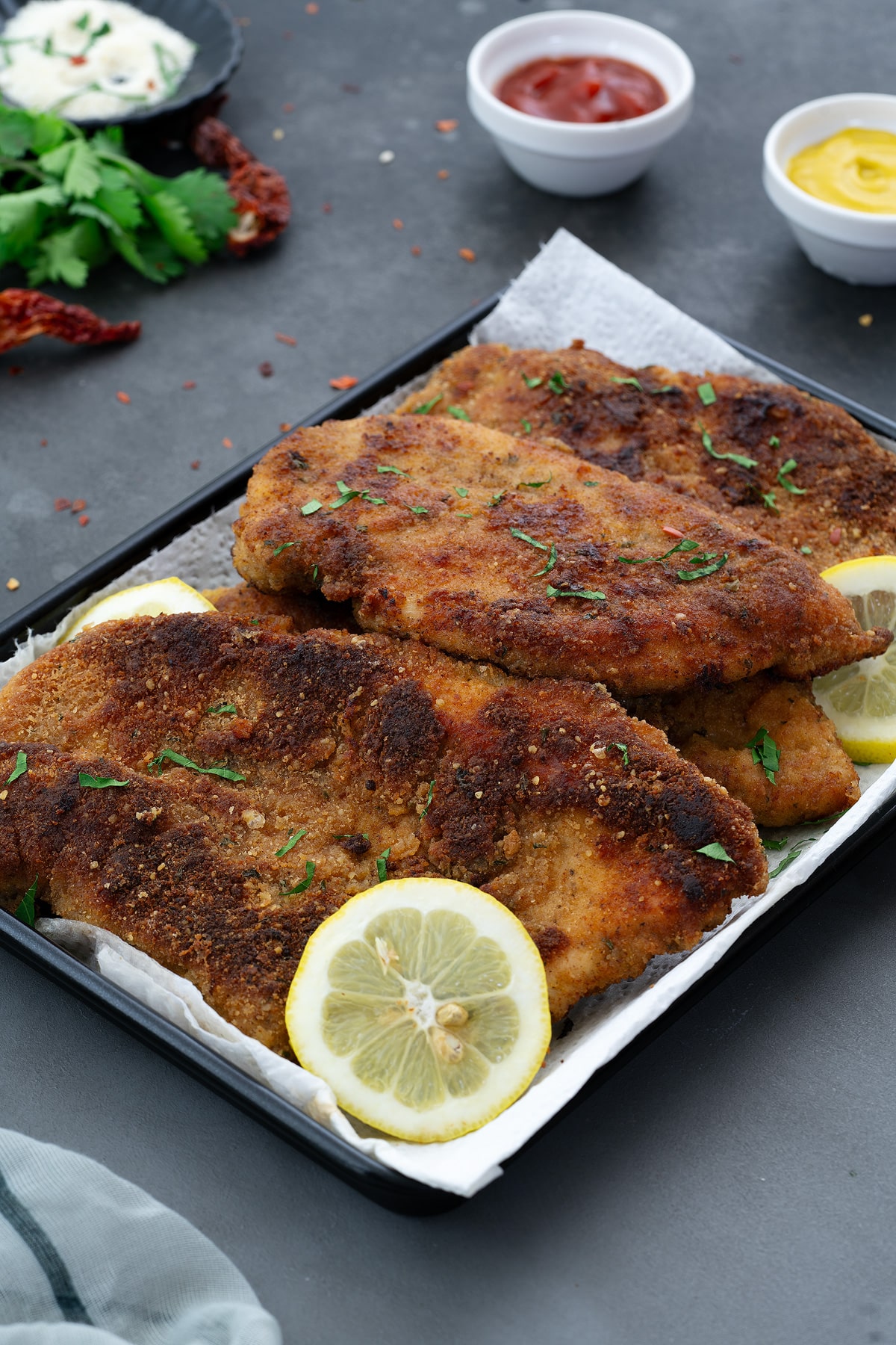 Crispy and Tender Chicken Cutlets on a black tray, presented on a grey table with fresh coriander leaves and zesty lemon slices. Accompanied by white cups of flavorful Ketchup and yellow mustard dips.