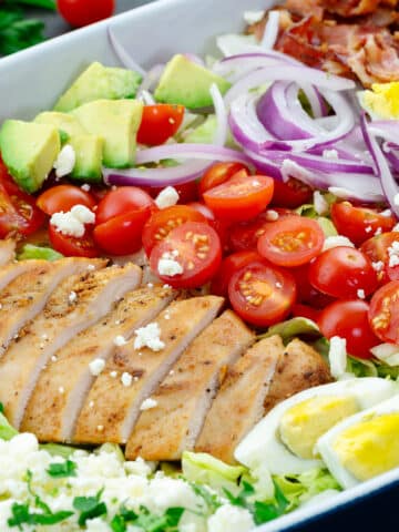 A delicious Cobb salad made with fresh iceberg lettuce, baked chicken breast, bacon, eggs, avocado, cherry tomatoes, ranch dressing, and feta cheese. The salad is beautifully arranged on a serving tray and is ready to be enjoyed.
