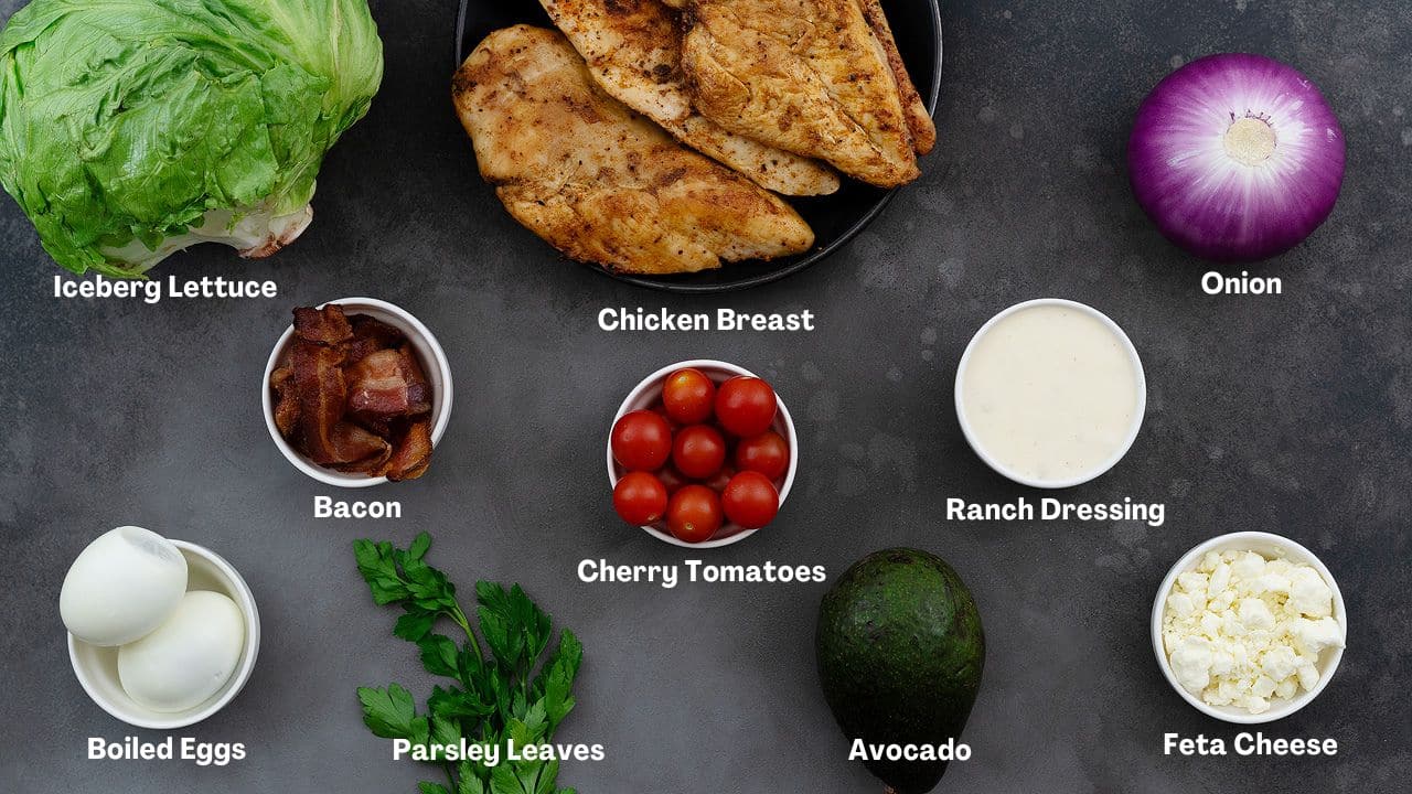 A variety of ingredients for a Cobb salad arranged on a grey table. The ingredients include iceberg lettuce, baked chicken breast, onion, bacon, cherry tomatoes, ranch dressing, boiled eggs, parsley leaves, avocado, and feta cheese.