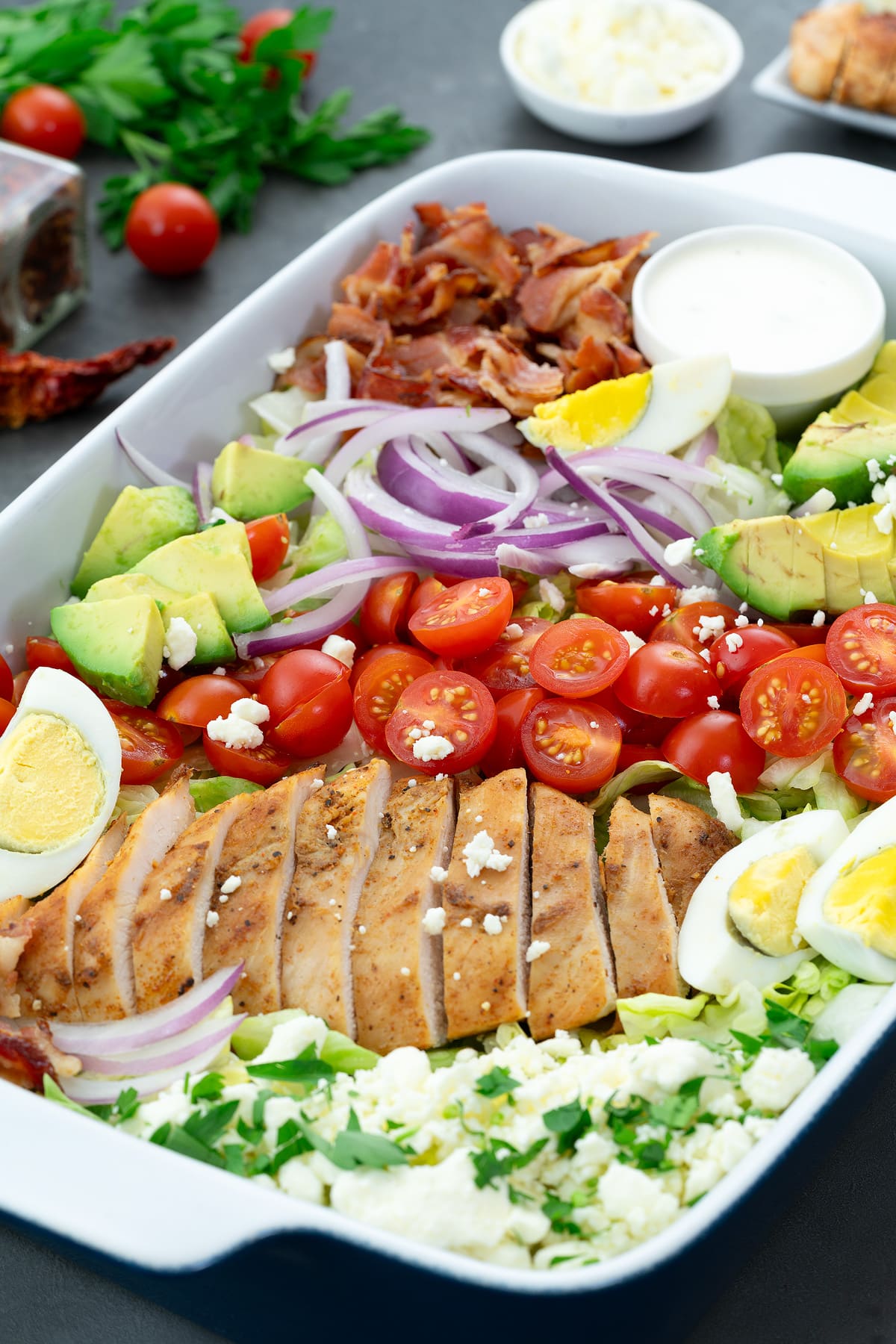 A delicious Cobb salad made with fresh iceberg lettuce, baked chicken breast, bacon, eggs, avocado, cherry tomatoes, ranch dressing, and feta cheese. The salad is beautifully arranged on a serving tray and is ready to be enjoyed.