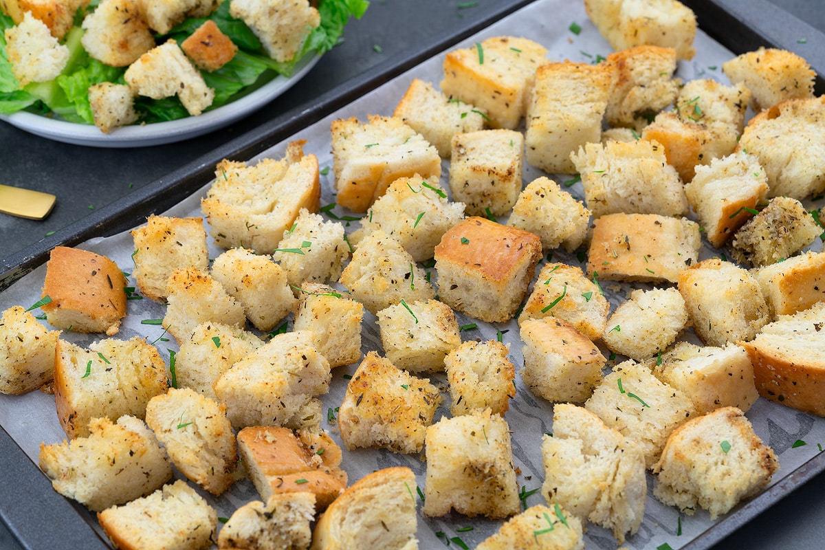 Homemade croutons on a baking tray, a small cup of pepper, and a plate of croutons and lettuce mix.