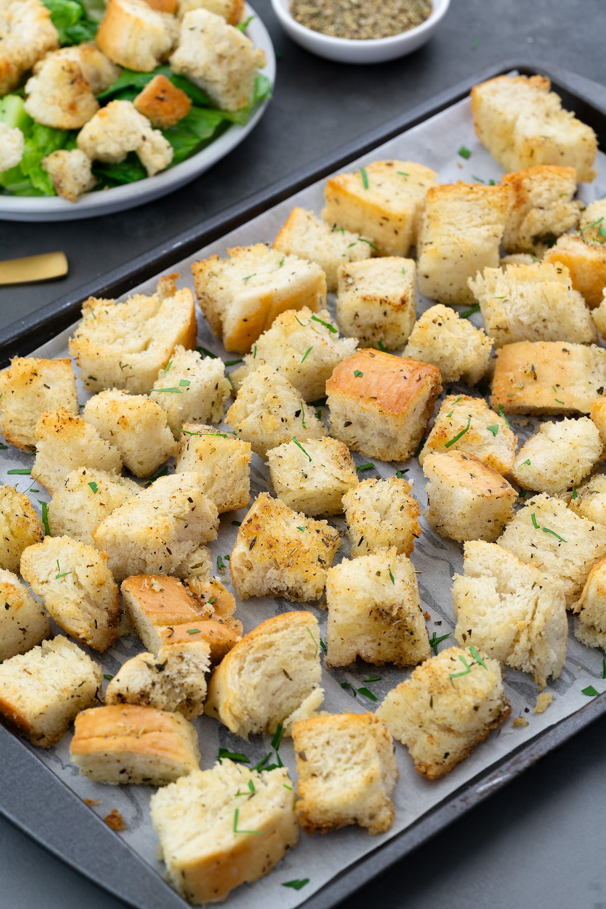Homemade croutons on a baking tray, a small cup of pepper, and a plate of croutons and lettuce mix.