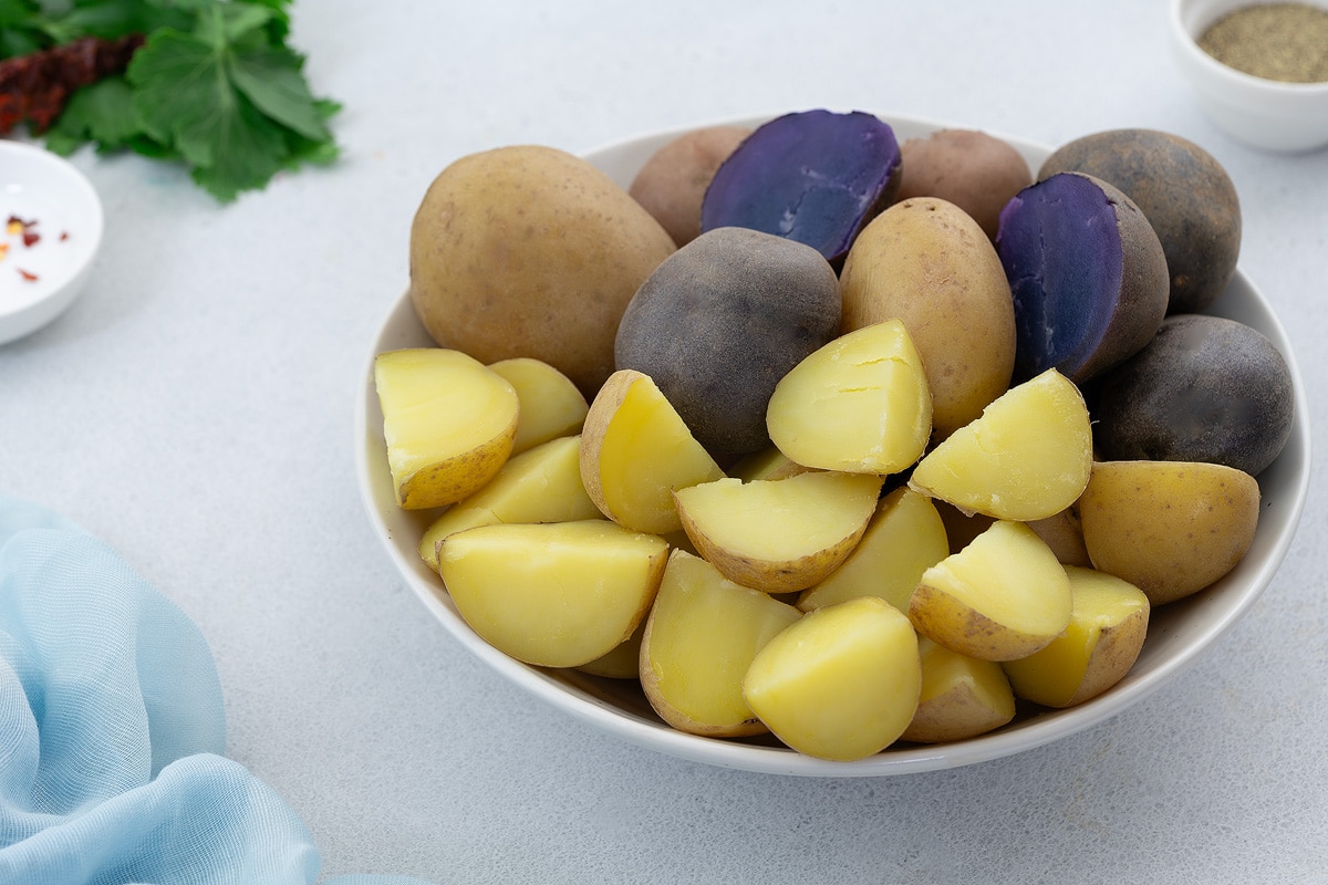 Assorted Boiled Potatoes in a White Bowl - Medley, Yukon Gold potatoes boiled whole and Honey Gold potatoes boiled and cubed, they are in purple, red and yellow colors. Accompanied by small bowls of salt and pepper.