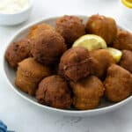 A bowl of golden brown hush puppies with lemon slices, accompanied by a cup of ketchup and tartar sauce. The hush puppies are resting on a white tabletop.