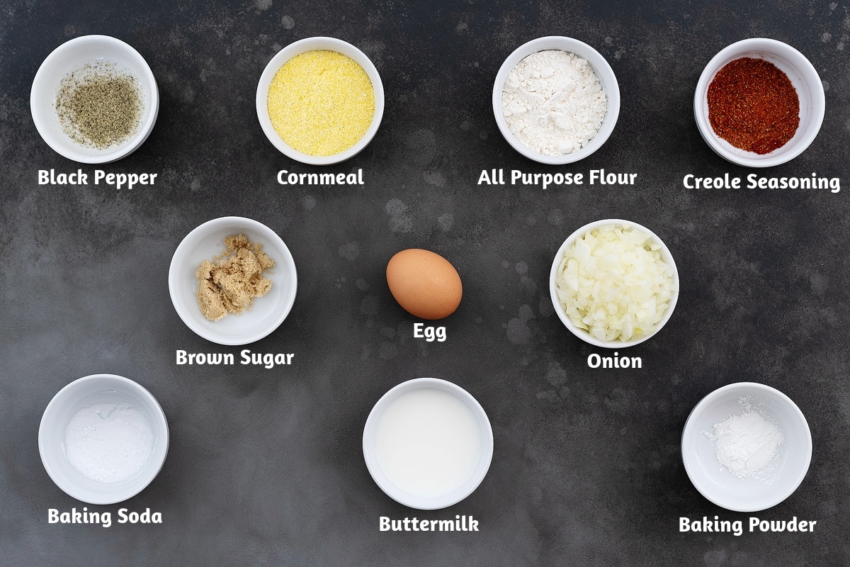 Ingredients for hush puppies, arranged on a grey table in white cups. The ingredients include black pepper powder, cornmeal, all-purpose flour, Creole seasoning, brown sugar, egg, onion, baking soda, buttermilk, and baking powder.